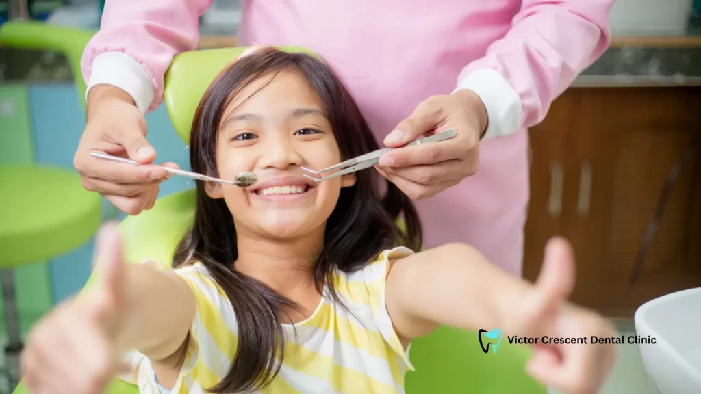 A happy young girl sits in the dental chair, giving a thumbs-up with a bright smile while a dental professional prepares instruments. This illustrates a successful and positive experience during a Berwick’s Smile Makeover or cosmetic consultation